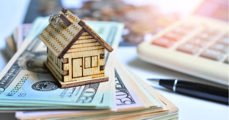 Wooden house model sitting on a stack of money with a pen and calculator in background
