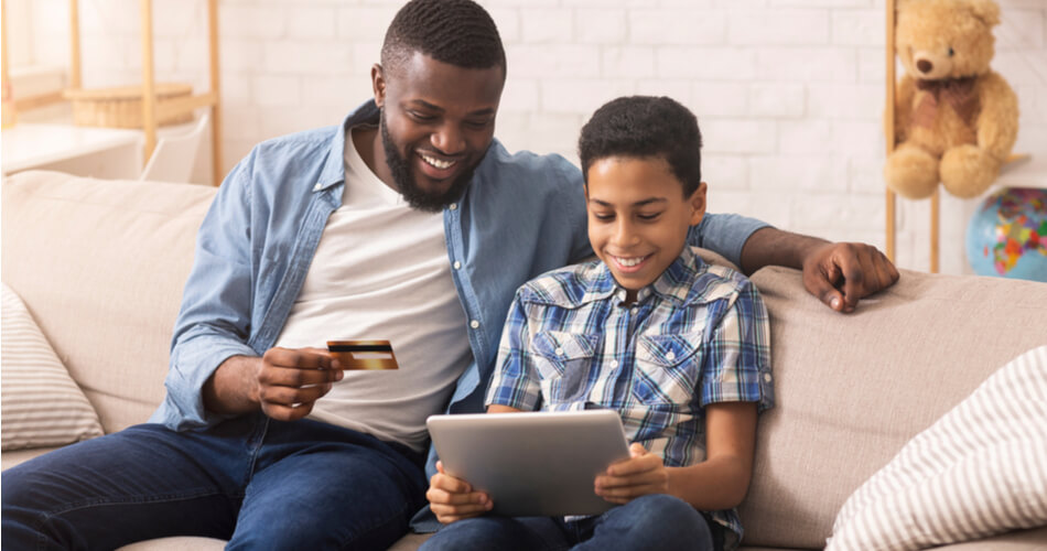 Father and son sitting on a couch smiling at a tablet while the father holds a credit card