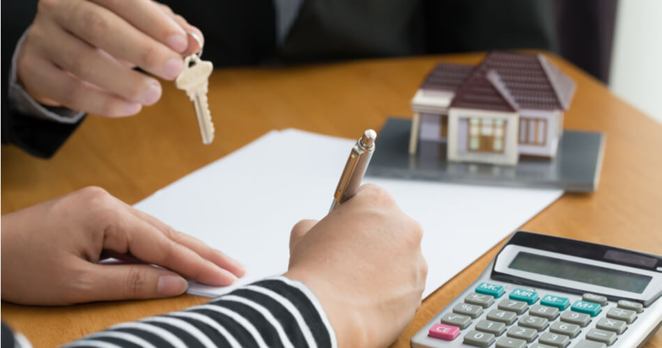 Woman signing purchase documents for a house with a model home and calculator on the table and a real estate agent handing house keys