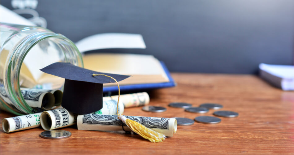 Glass jar of coins and rolled dollar bills with a small graduation cap on top and an open book in the background