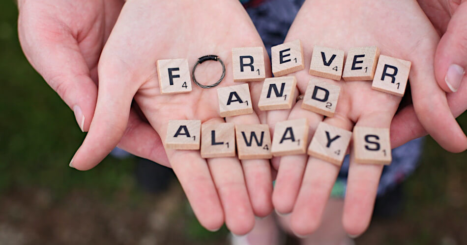 hands of couple hold scrabble pieces spelling forever and always with ring in place of letter o