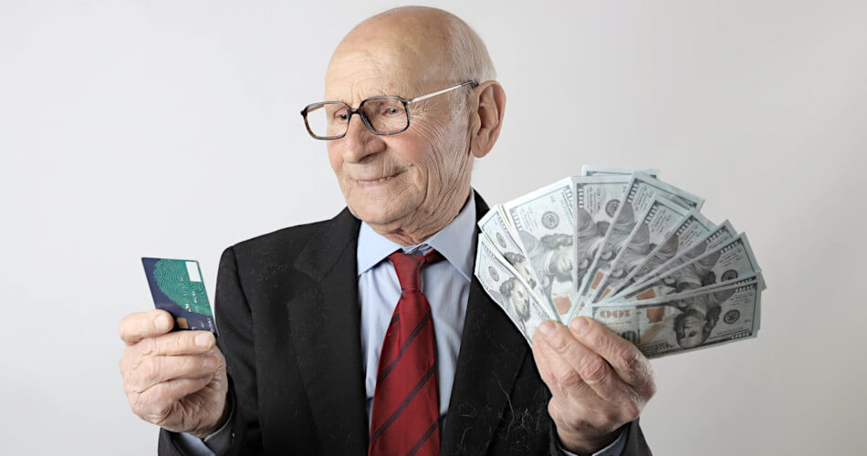 smiling elderly man holds credit card in one hand, several $100 bills in the other
