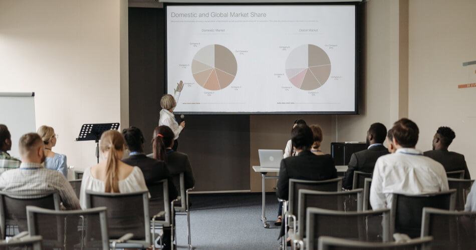 The instructor in a business course shows Domestic and Global Market Share pie graphs as an example of how business data can be presented in a visually engaging way. A business course like this might well qualify for your employer's tuition reimbursement program.